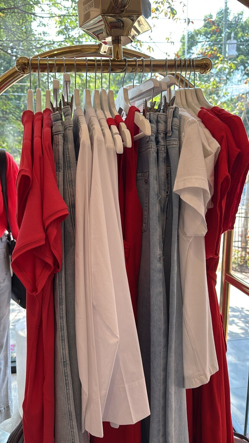 A brass luggage-cart rack of red tops and dresses, white shirts and light denim jeans, seen through a shop window