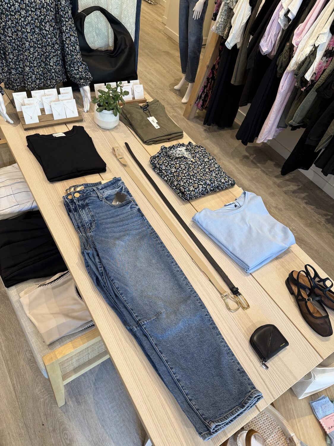 A pale wood table with a pair of jeans laid out beside belts, a black top, a floral blouse, a powder blue tee and shoes